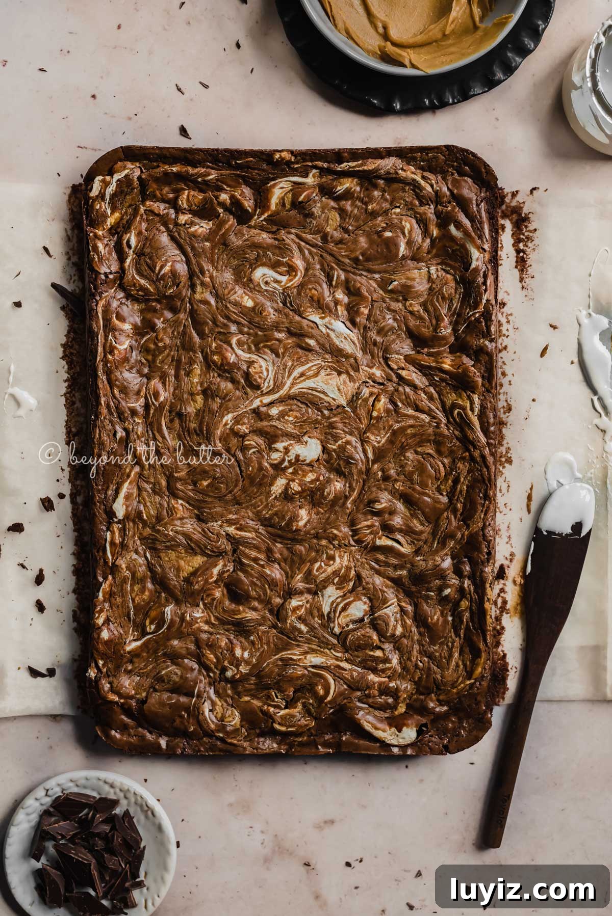 Marshmallow Peanut Butter Brownies Recipe — Gooey Chocolate Bars 7 Uncut marshmallow peanut butter brownies on parchment paper and bowl of marshmallow fluff and spreader tool beside it | All Images © Beyond the Butter™