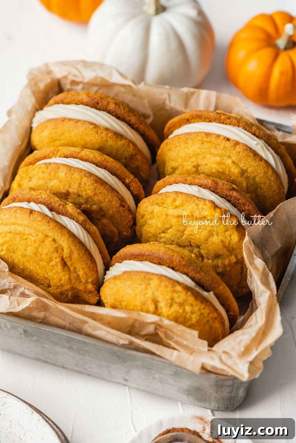 Pumpkin Whoopie Pies Recipe: Soft Spiced Sandwich Cookies 2 Six pumpkin whoopie pies placed in a brown parchment paper lined metal baking tin with mini orange and white pumpkins placed around it on a white background.