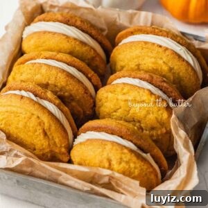 Pumpkin Whoopie Pies Recipe: Soft Spiced Sandwich Cookies 22 Six pumpkin whoopie pies placed in a brown parchment paper lined metal baking tin with mini orange and white pumpkins placed around it on a white background.