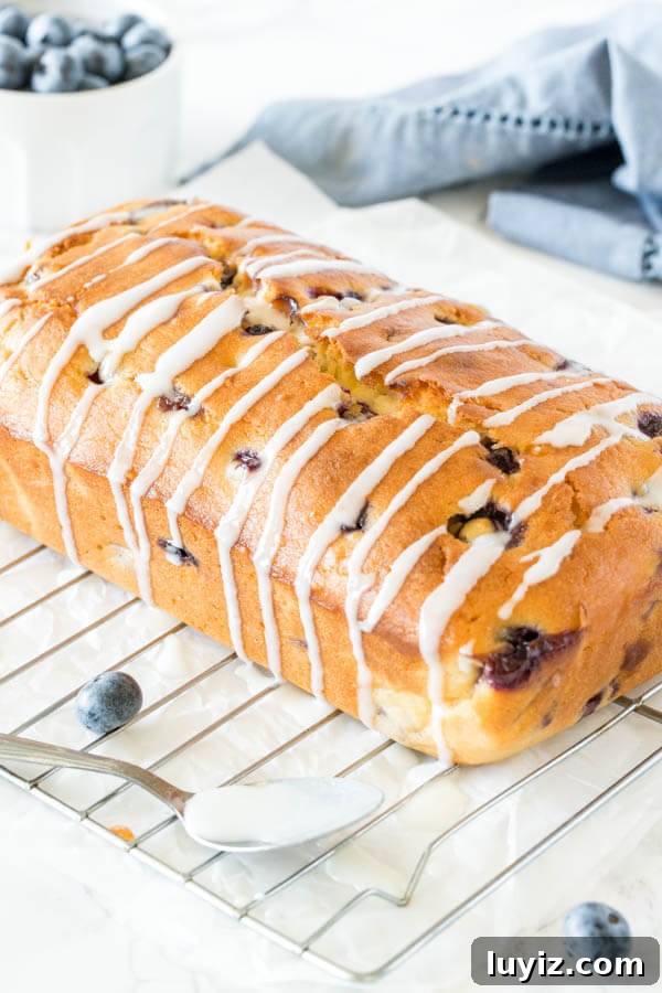 Moist Blueberry Loaf Recipe with Lemon Zest 3 Blueberry Bread drizzled with vanilla glaze on a cooling rack with a small bowl of blueberries in the background.