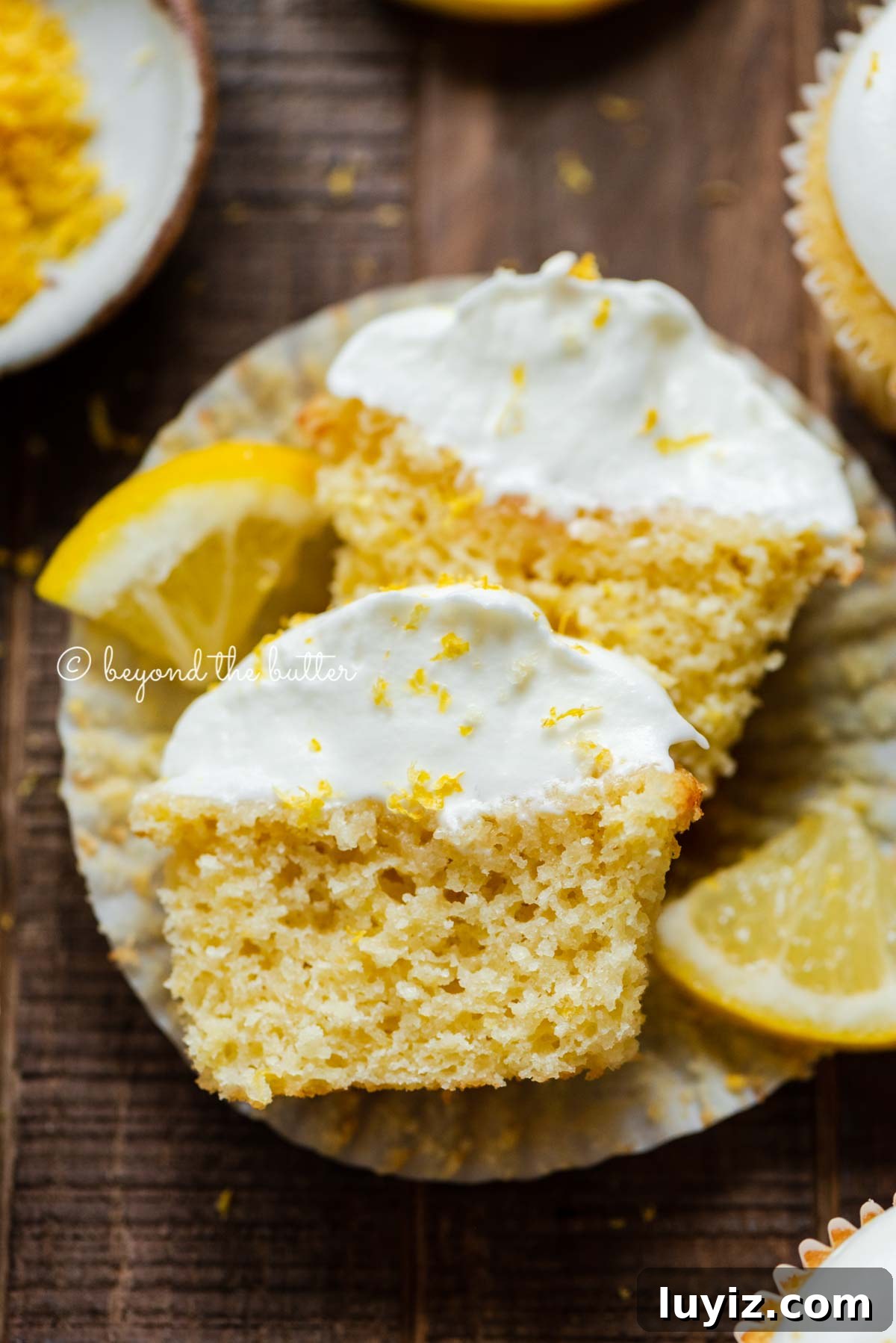 Mini Lemon Cupcakes: Perfect Small-Batch Recipe for 6-8 Servings 2 Sliced open lemon cupcake with whipped lemon cream cheese frosting on cupcake liner and 2 wedges of lemons on a dark wood background