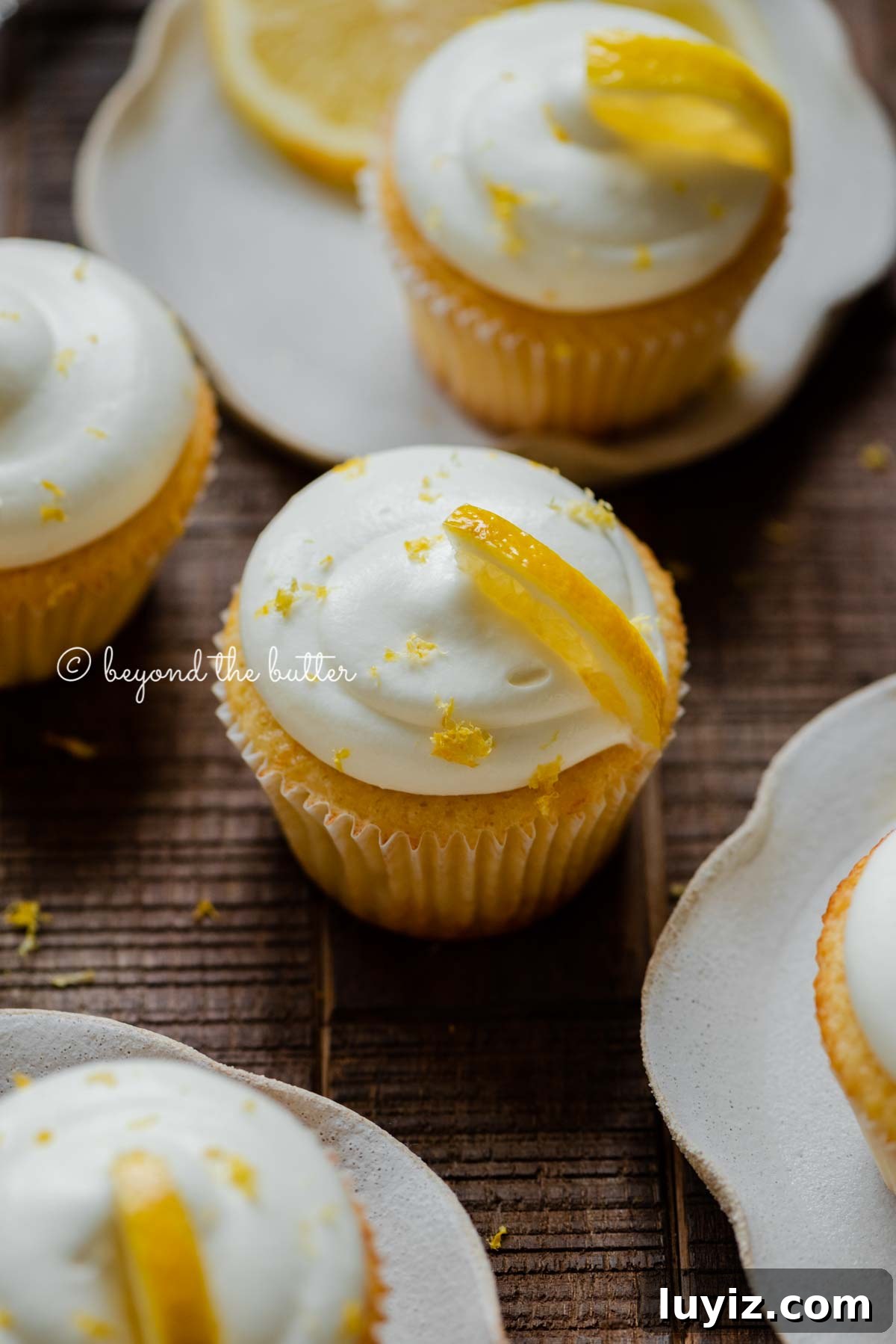 Mini Lemon Cupcakes: Perfect Small-Batch Recipe for 6-8 Servings 3 Randomly placed lemon cupcakes with lemon cream cheese frosting, garnished with lemon triangles with a small bowl of lemon zest on a dark wood background