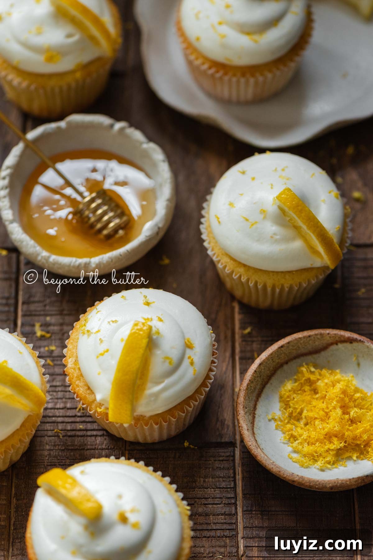 Mini Lemon Cupcakes: Perfect Small-Batch Recipe for 6-8 Servings 8 Randomly placed lemon cupcakes with a small bowl of honey and small bowl of lemon zest on a dark wood background