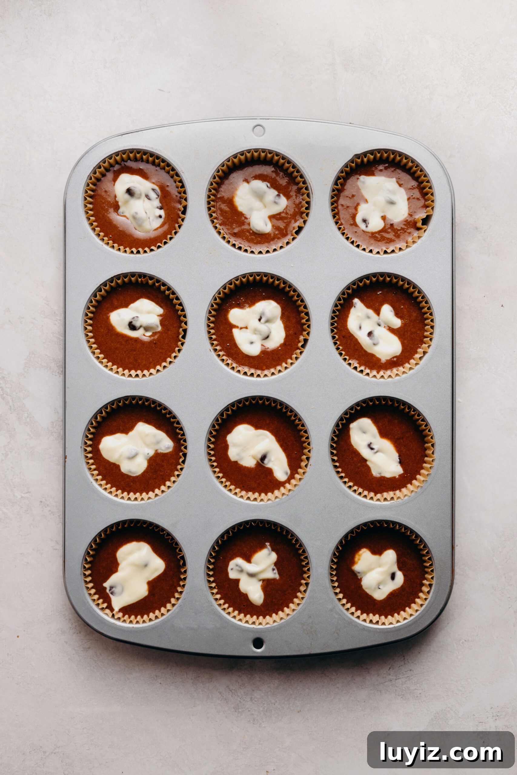 Decadent Black Bottom Cupcakes with Cream Cheese Swirl 11 Metal cupcake tin filled with chocolate cupcake batter topped with chocolate chip cream cheese filling on a white marbled background.