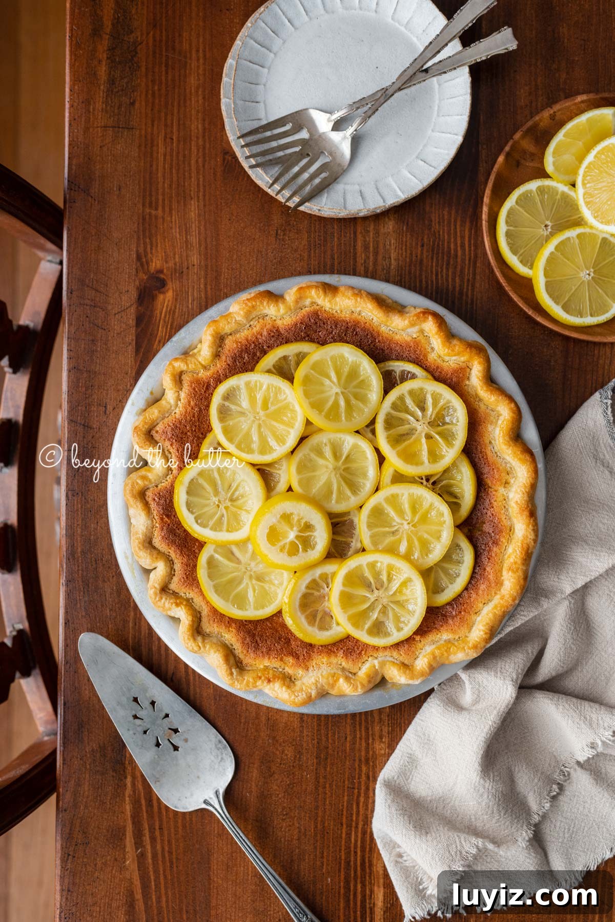 Classic Lemon Sponge Pie Recipe — Light, Zesty Dessert 3 lemon sponge pie decorated with candied lemon sliced on a dark wood background with small dessert plates, napking, pie server, and small plate of sliced lemons | All images © Beyond the Butter®