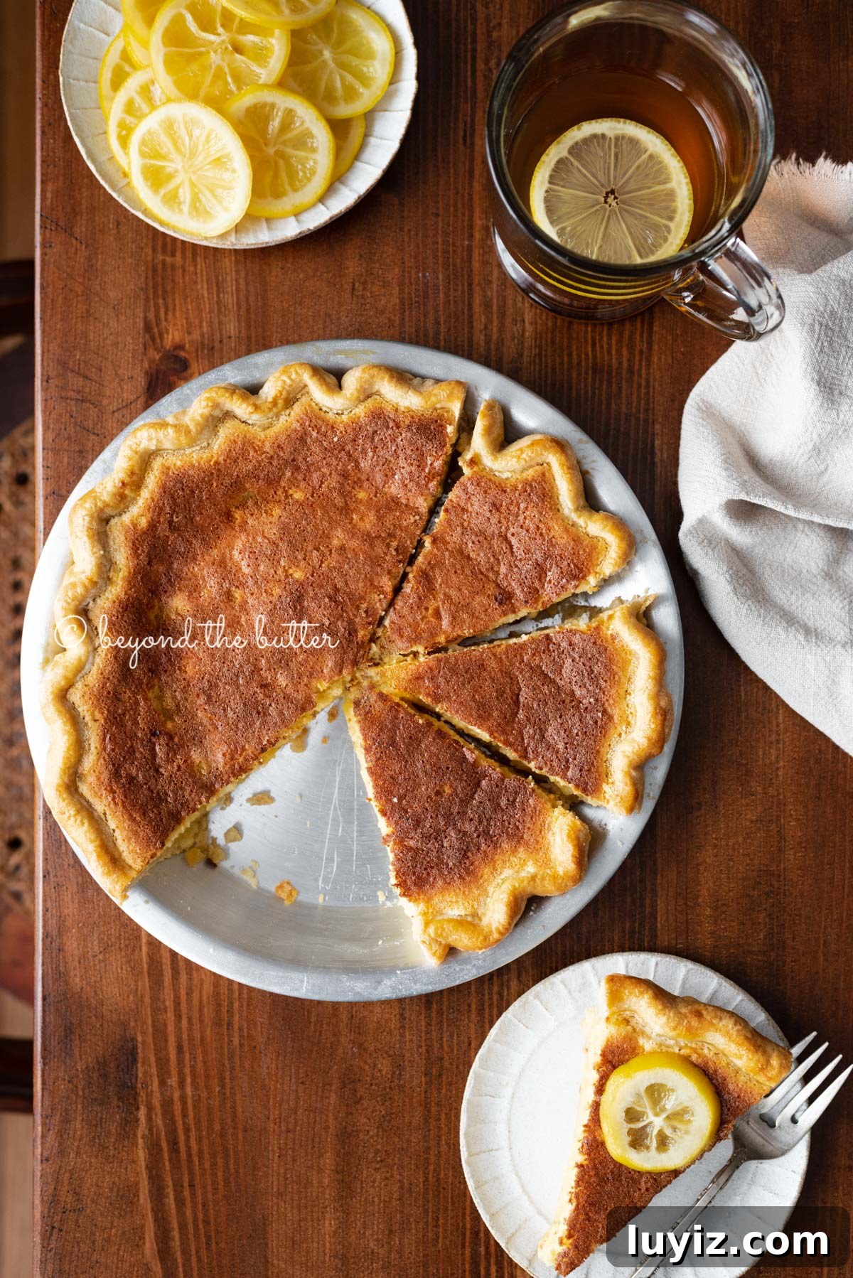 Classic Lemon Sponge Pie Recipe — Light, Zesty Dessert 6 Lemon sponge pie with cut slices on dark wood background with hot lemon tea, small plate of sliced lemons, and slice to the side | All images © Beyond the Butter®