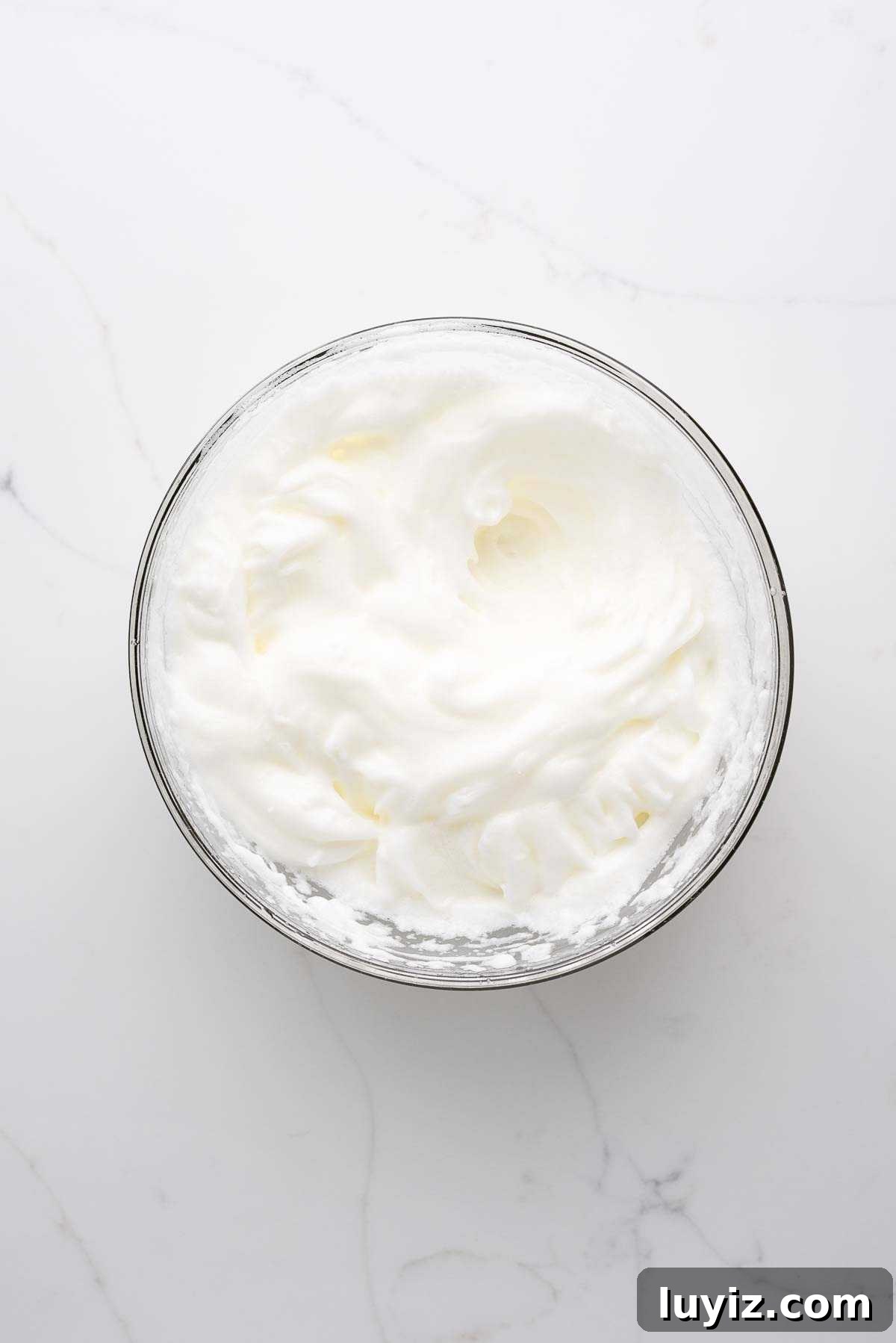 Egg whites whisked to stiff peaks in a small glass bowl on a white marbled background.