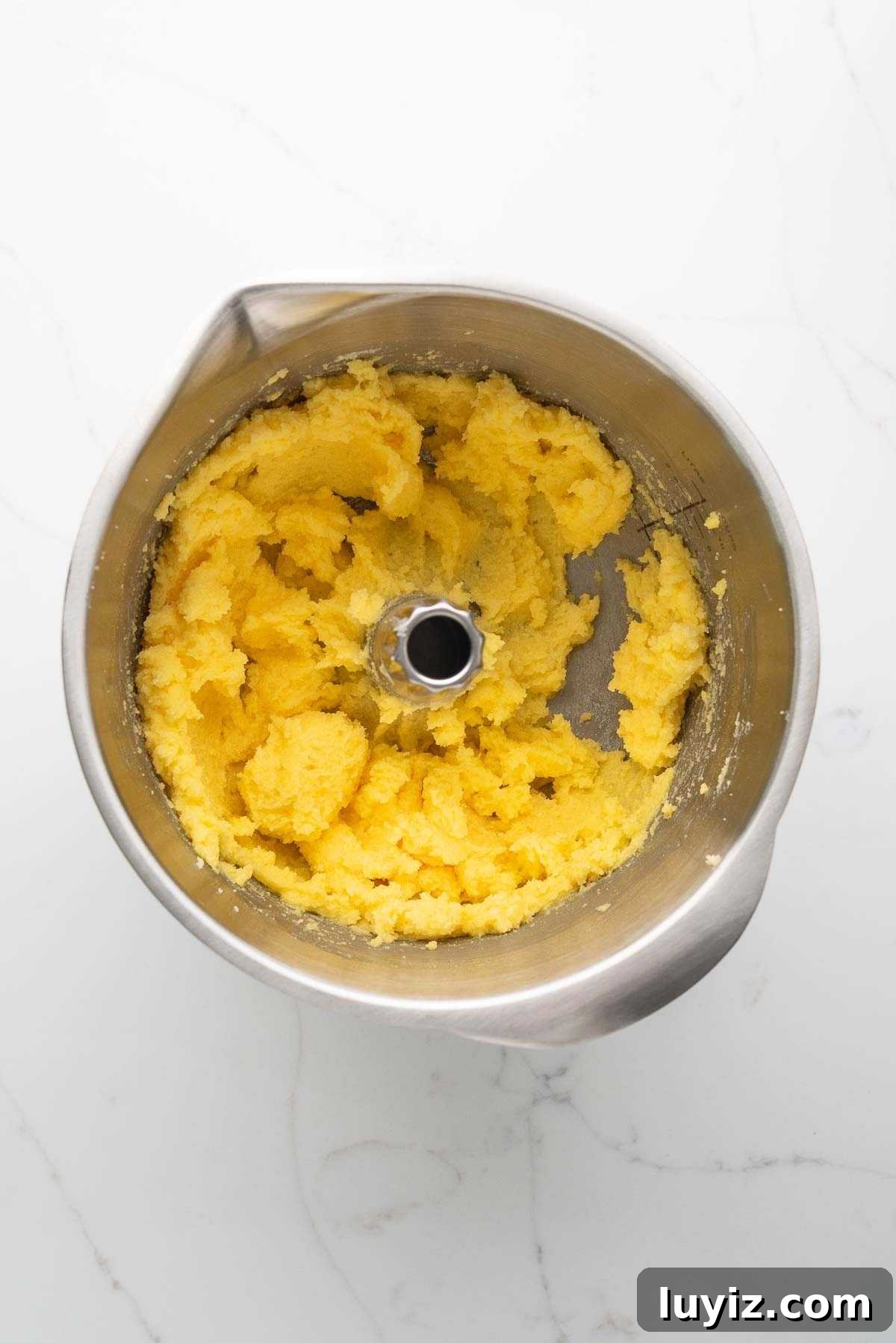 Beaten sugar, butter, and egg yolks in a stainless steel mixing bowl on a white marbled background.