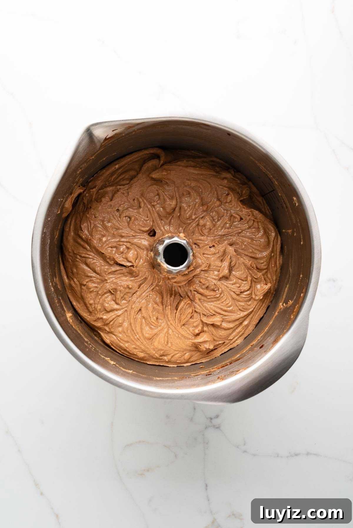 Flour mixture and buttermilk added to the German chocolate cake batter in a stainless steel mixing bowl on a white marbled background.