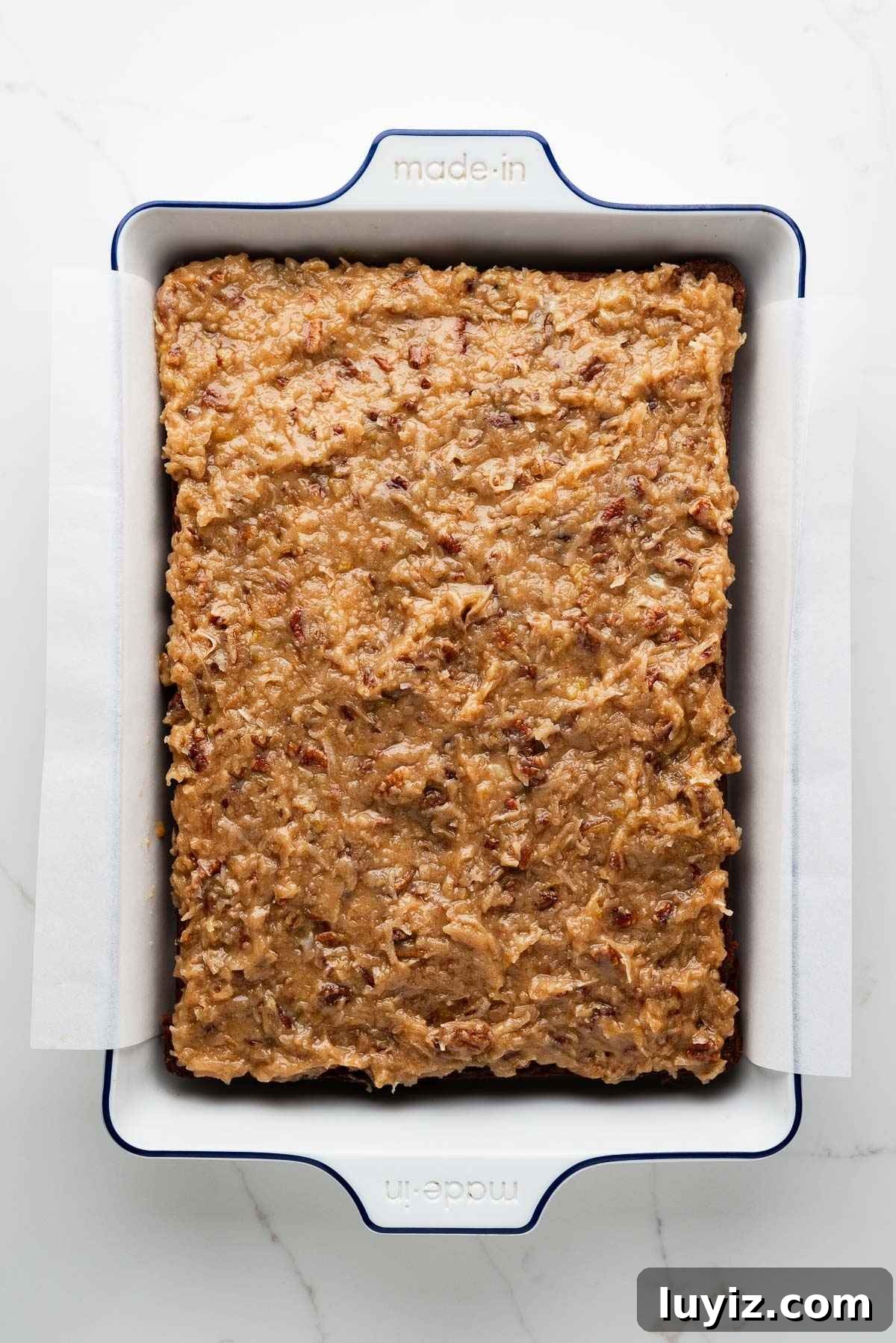 Coconut pecan frosted German chocolate sheet cake in a parchment paper lined white ceramic baking dish on a white marbled background.
