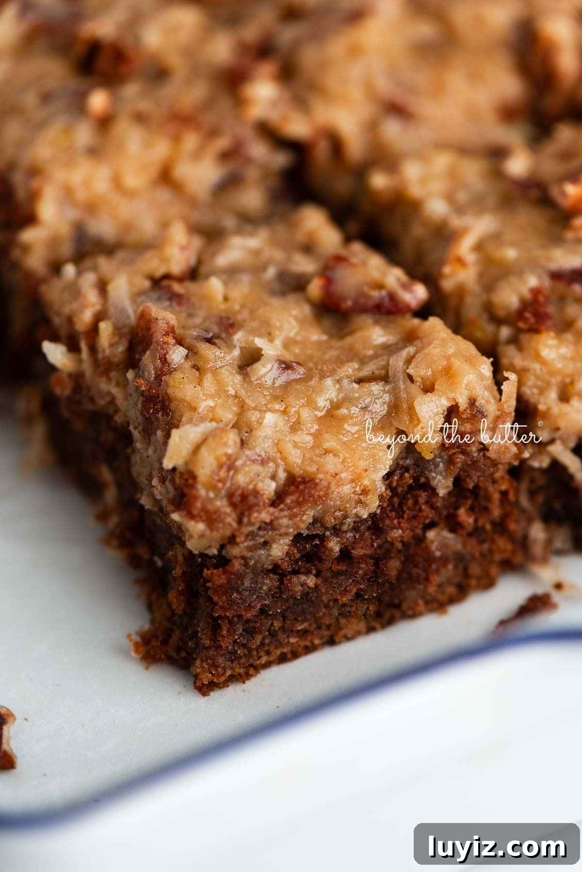 Sliced German chocolate cake with coconut pecan frosting in a parchment paper lined ceramic baking pan.