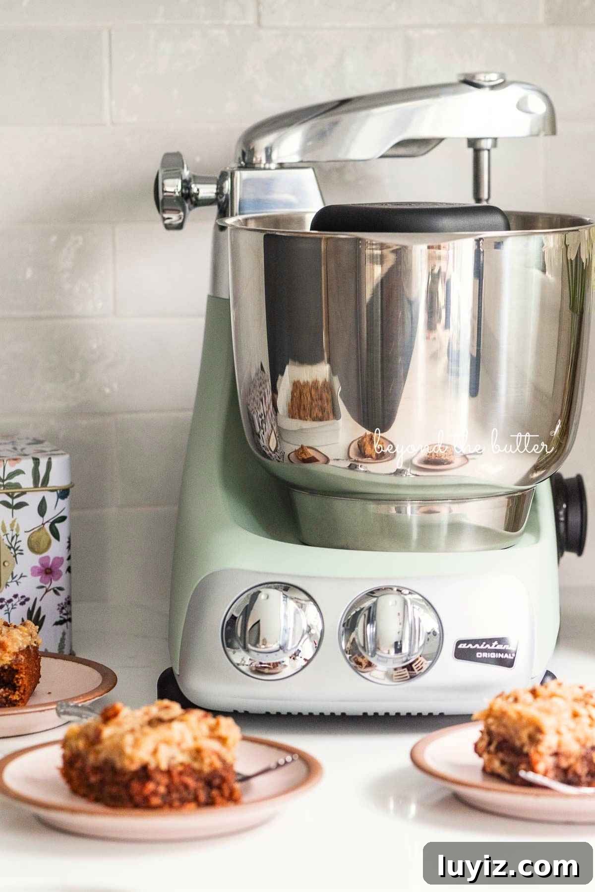 Ankarsrum assistent original mixer in nordic sage on white quartz kitchen counter with subway tile backsplash with recipe box and dessert plates with german chocolate sheet cake around it.