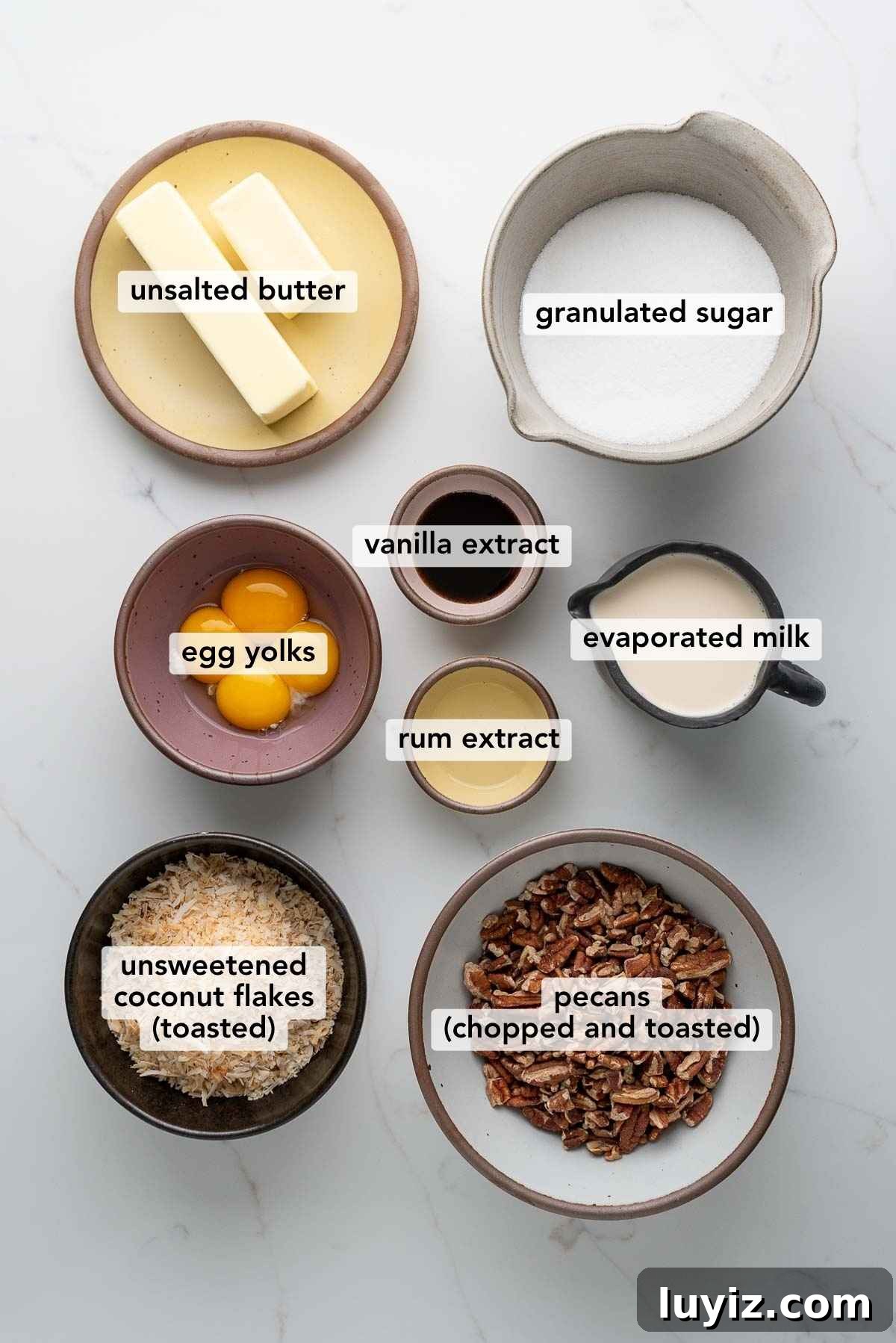 Ingredients in various size bowls labeled as unsalted butter, granulated sugar, egg yolks, vanilla extract, rum extract, evaporated milk, unsweetened coconut flakes (toasted), and pecans (chopped and toasted) on a white marbled background.