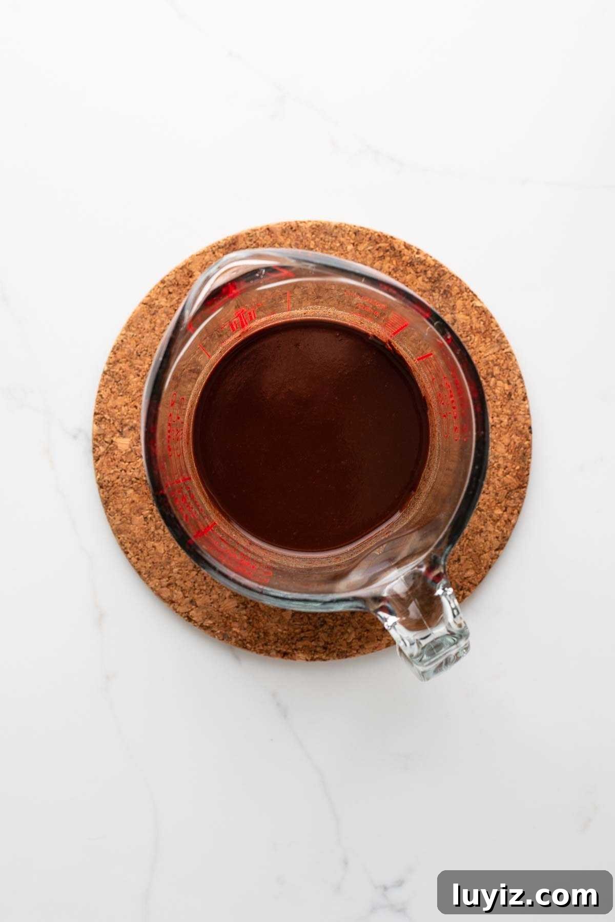 Melted chocolate and water together in a glass measuring cup sitting on a cork trivet on a white marbled background.