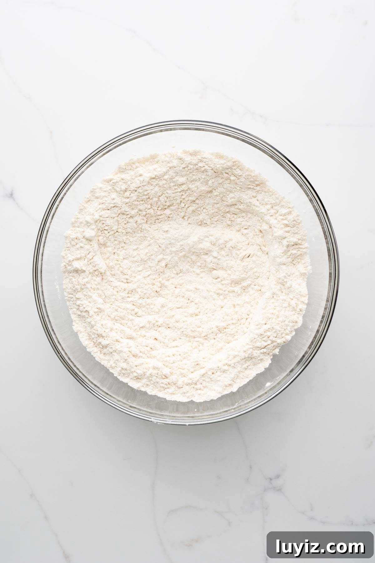 Whisked all purpose flour, baking soda, baking powder, and salt in a medium-sized glass bowl on a white marbled background.