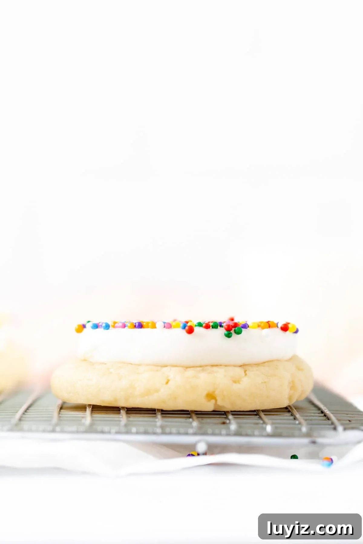 Dot cake cookie on a cooling rack with a thick layer of vanilla frosting and sprinkles on top