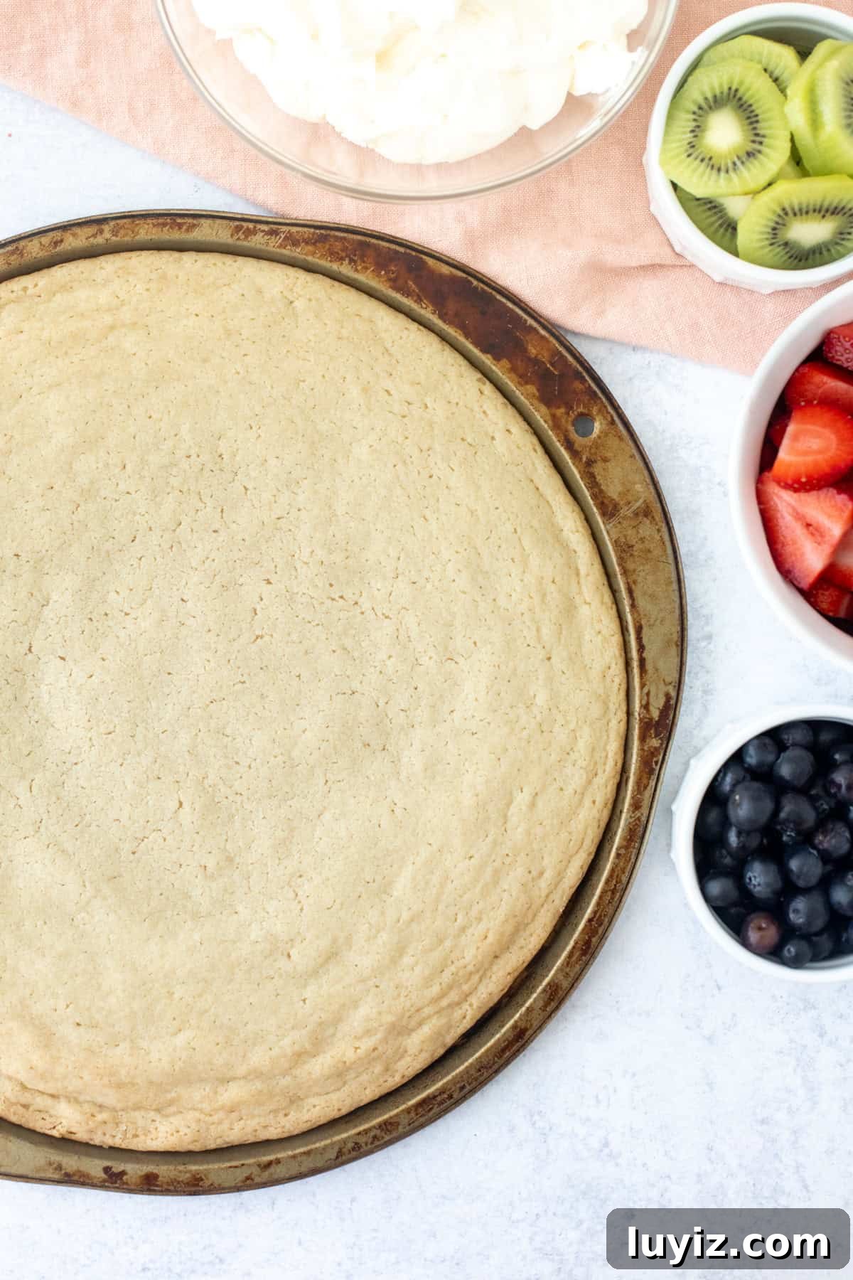 Sugar cookie made in a large pizza pan with bowl of whipped cream cheese frosting and bowls of fruit